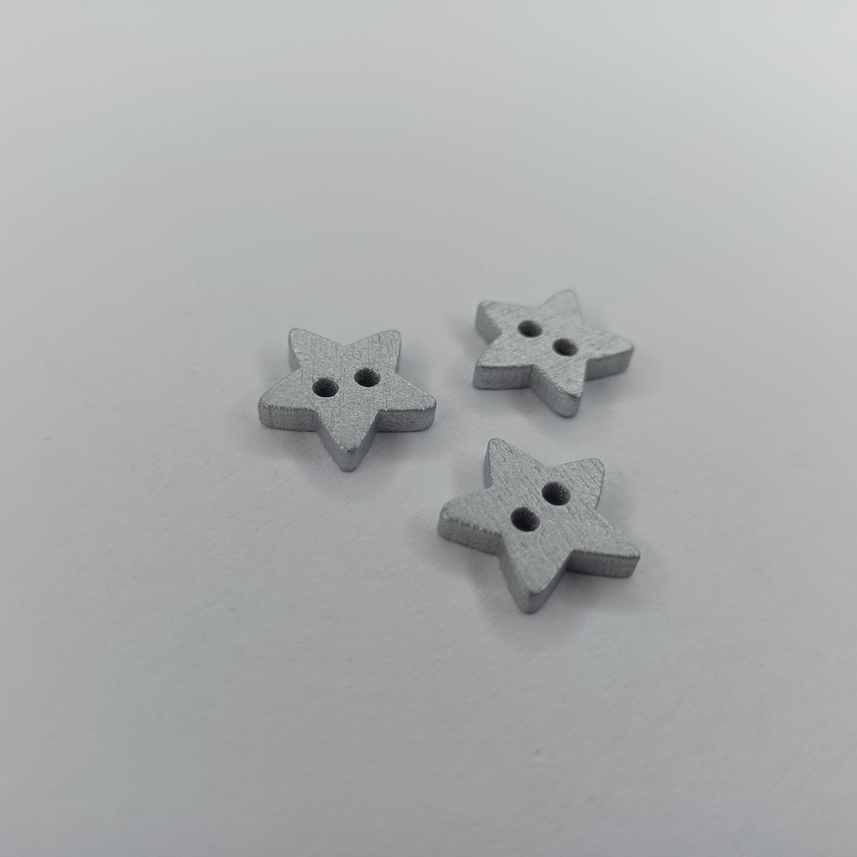 Star Wood Buttons 13mm 2 Holes – Silver & Gold – Three small star-shaped buttons with two holes, finished in a textured light gray, arranged on a plain light gray background.